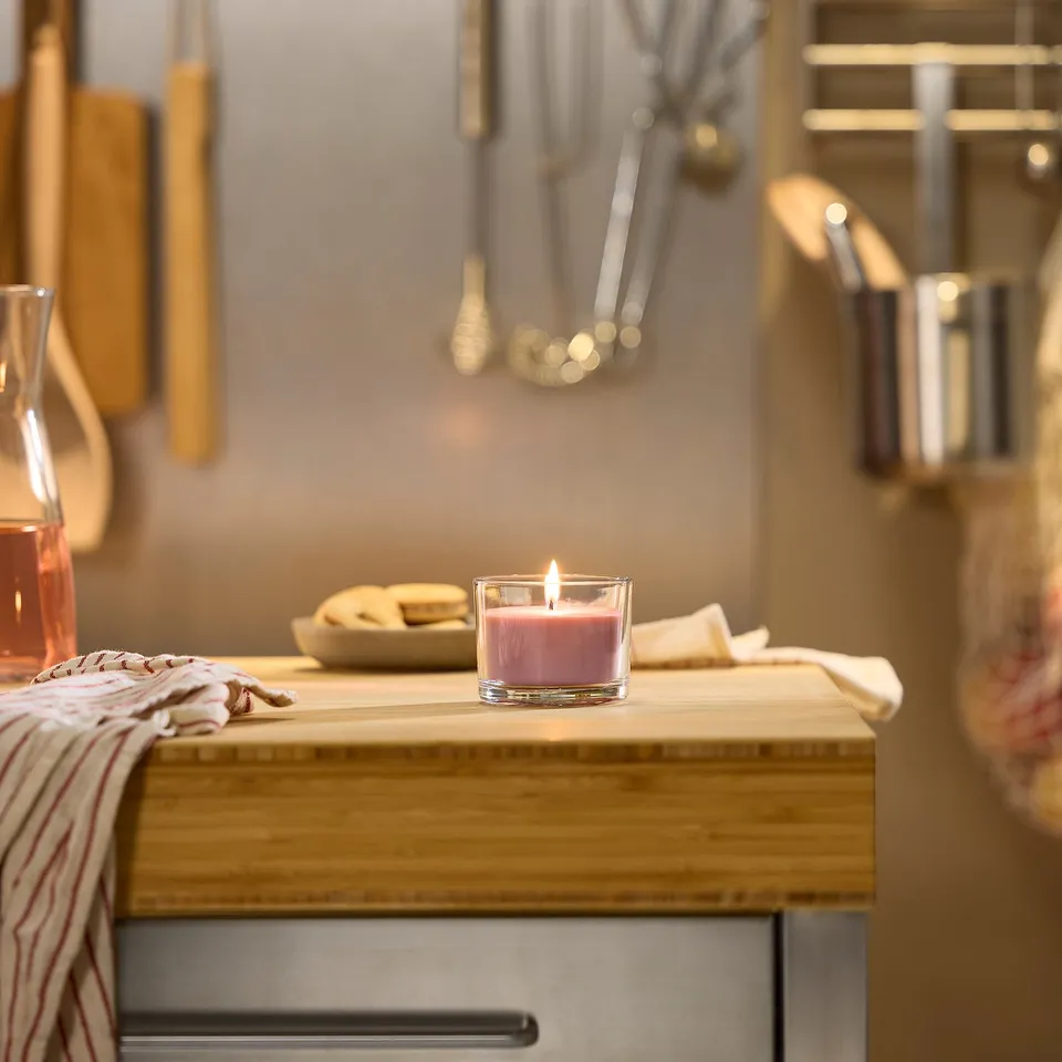 Small pink scented candle in a glass cup on a wooden shelf with cooking utensils.
