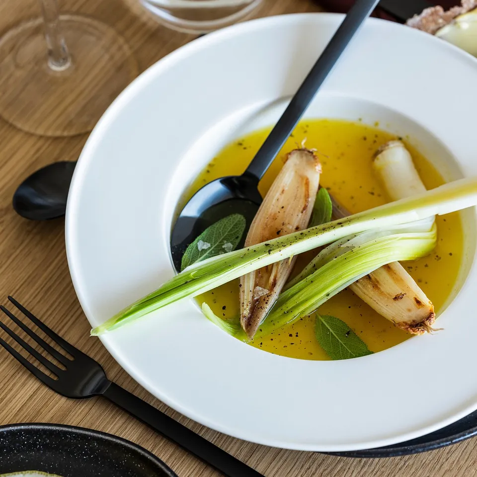 White deep plate with grilled leeks in yellow sauce, black spoon and fork on wood table.