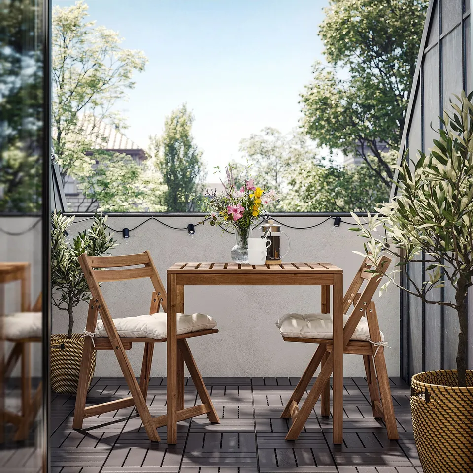 Small wooden balcony table set with cushioned chairs, plant, vase, and lights.