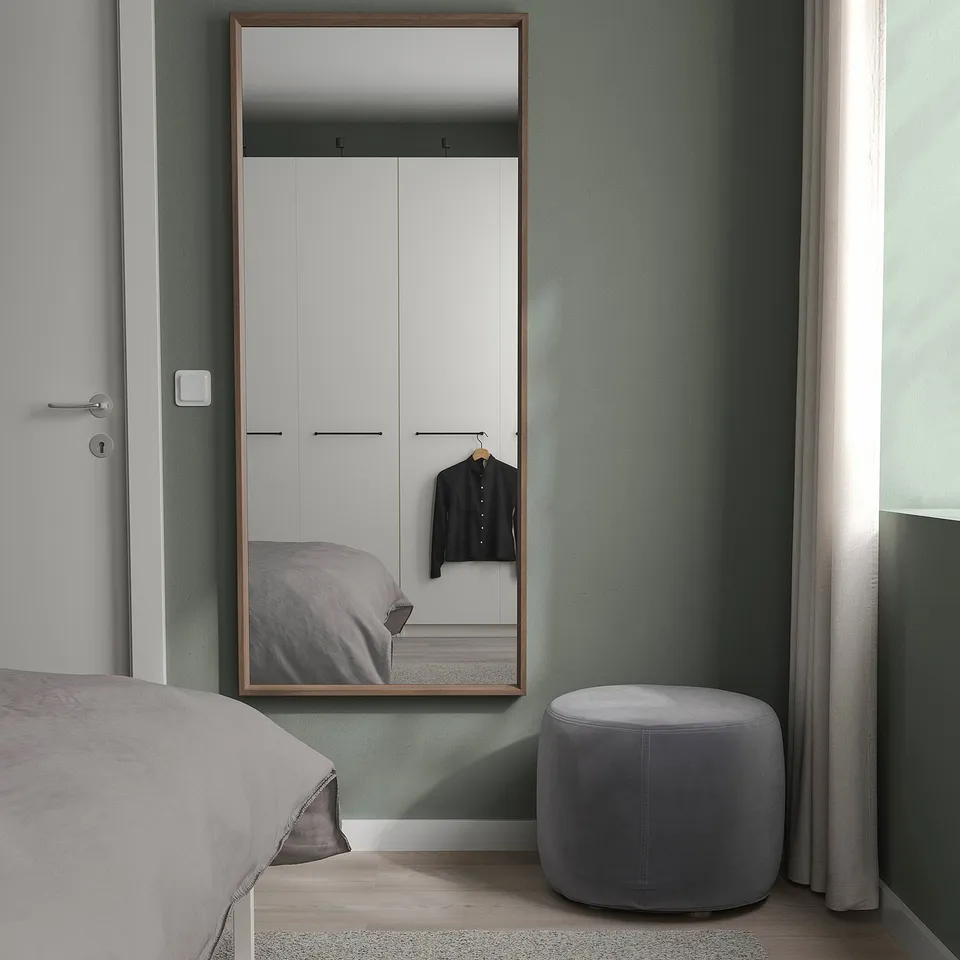 Modern bedroom with NISSEDAL full-length mirror - wooden frame, reflecting wardrobe and bed; grey ottoman beside.