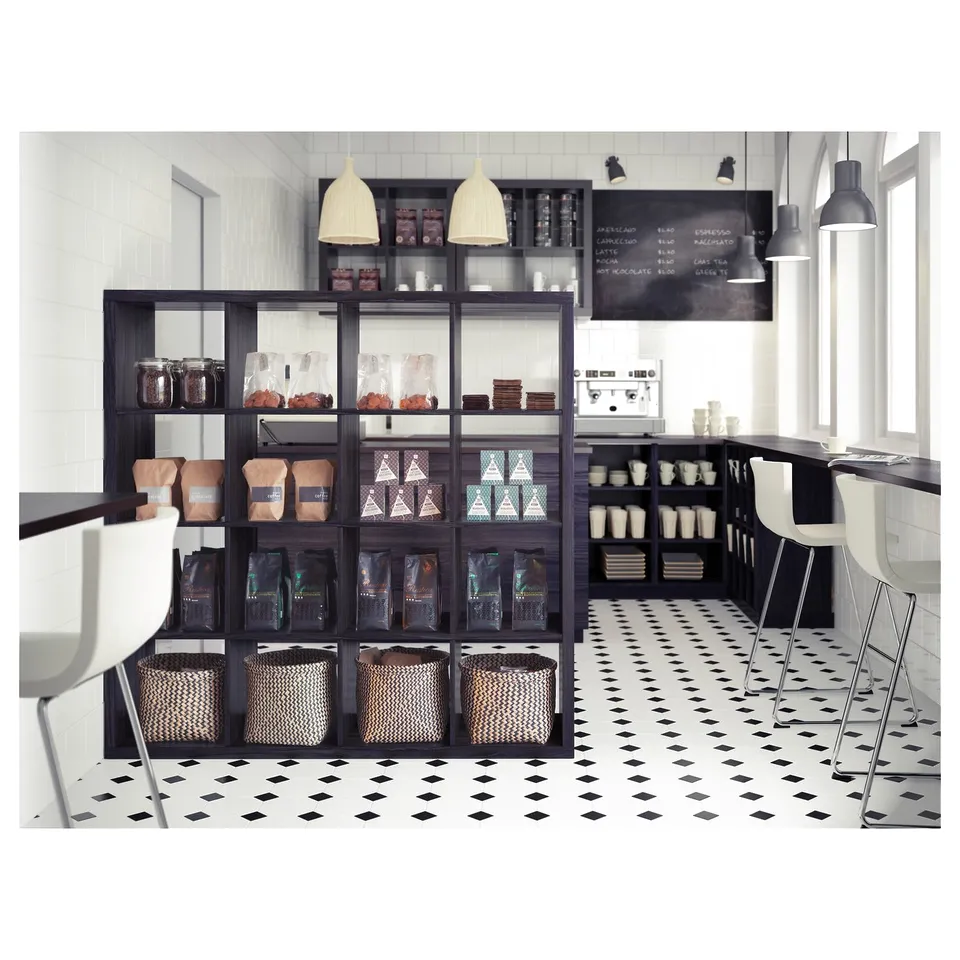 Modern coffee shop with KALLAX shelves displaying bags of coffee and pastries, black and white tiles, white chairs, and black counter.