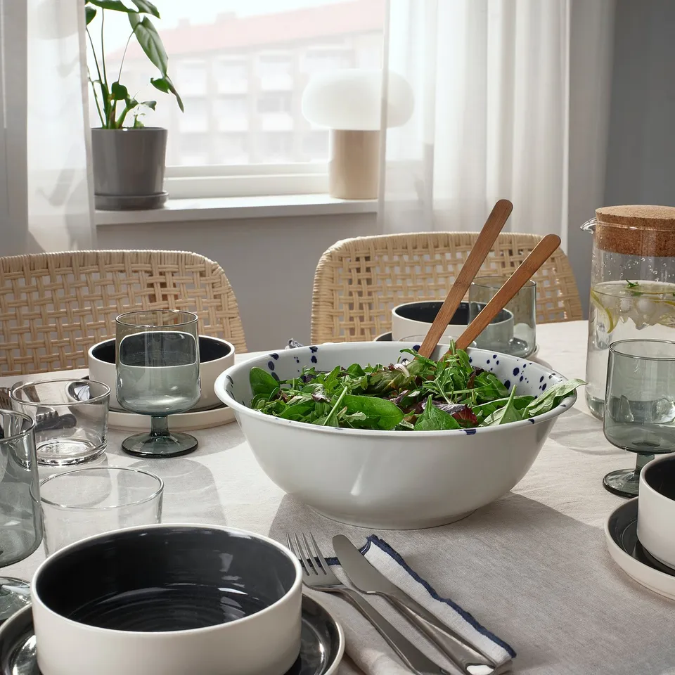 A dining table set with a salad bowl, glasses, cups, cutlery, and wicker chairs. Sunlight streams through the window.