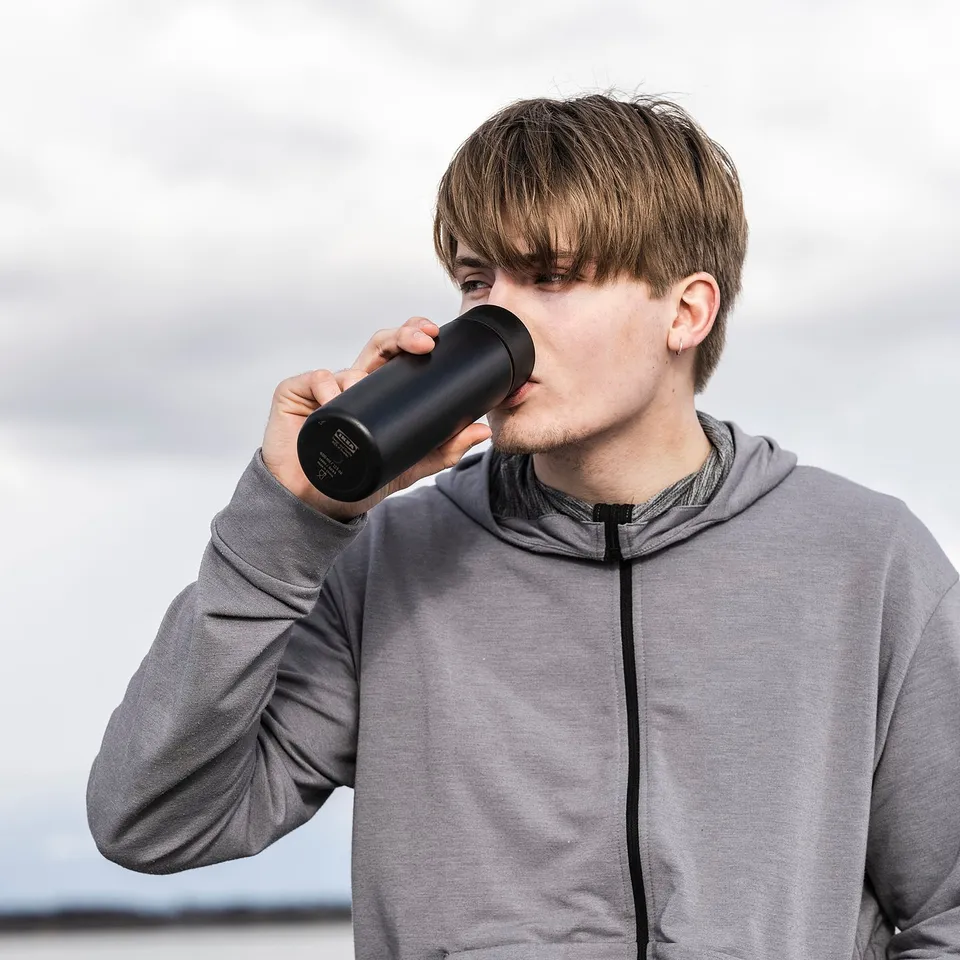 A person drinks from a black, cylindrical, stainless steel UNDERSÖKA travel mug outdoors, wearing a grey hoodie.