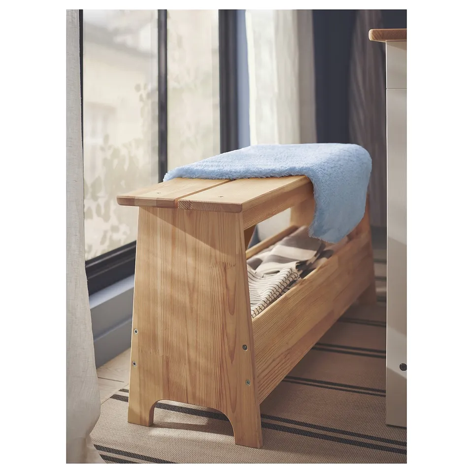 Light wooden bench with a blue, soft blanket draped over it, placed on a striped rug next to a window.