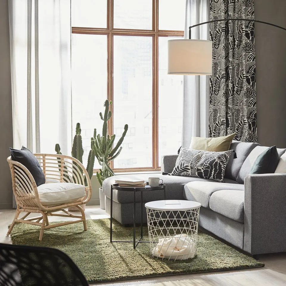 Modern living room with grey sofa, black coffee table, white side table, black lamp, wicker chair, large window, green rug, and black cabinet.