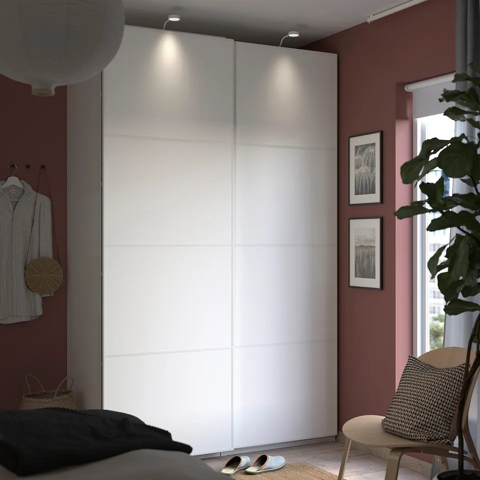 Modern bedroom with PAX white wardrobe, red wall, plant, and wooden chair.