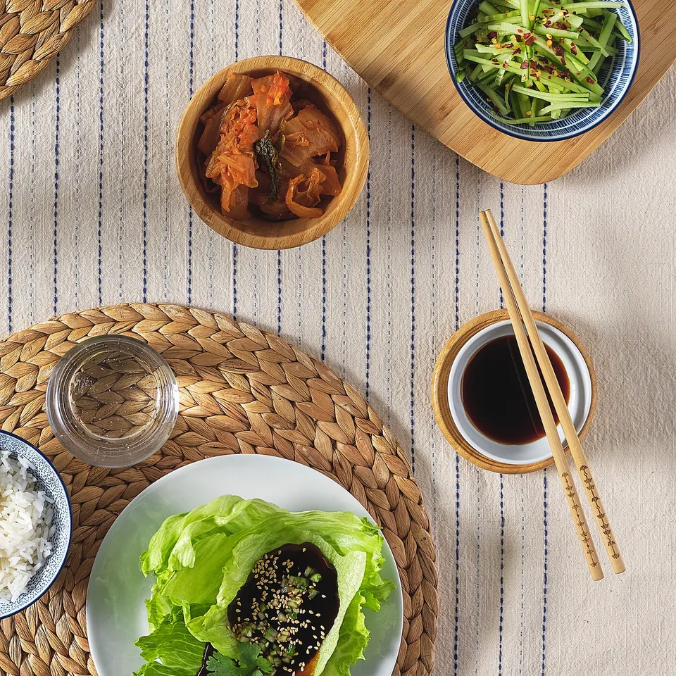 A meal setup with wooden bowls, chopsticks, and plates; includes lettuce wraps, rice, kimchi, and cucumber salad on a striped tablecloth.