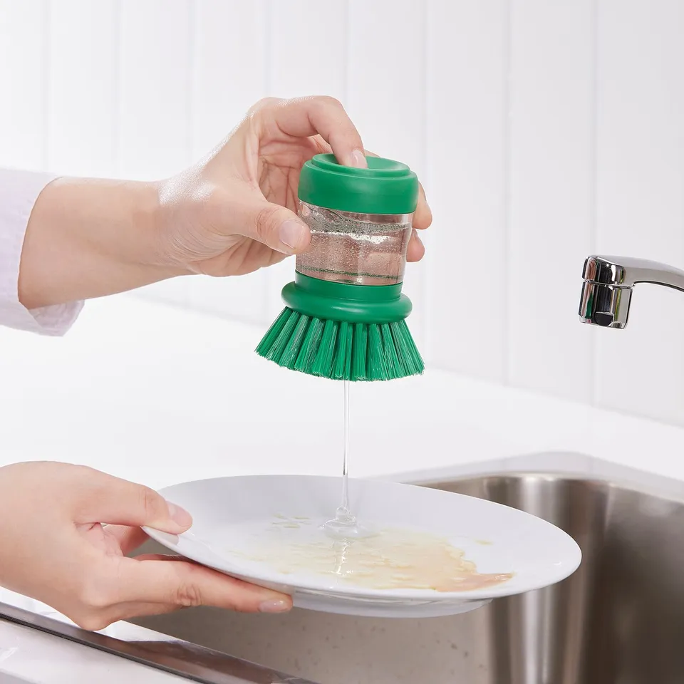 Person presses green brush, dispensing soap onto a dirty plate in a sink.
