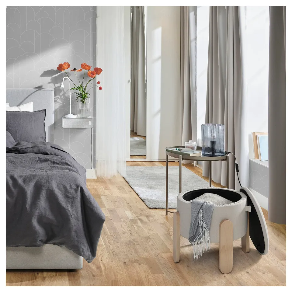 Modern bedroom with FÖRLUNDA pouffe in grey fabric, light wood legs, near bed with dark blanket, flower vase, grey curtains.