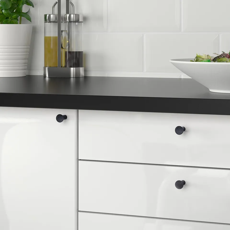 Modern kitchen with white cabinetry and black handles; a bowl of salad sits on a black countertop against a white brick backsplash.