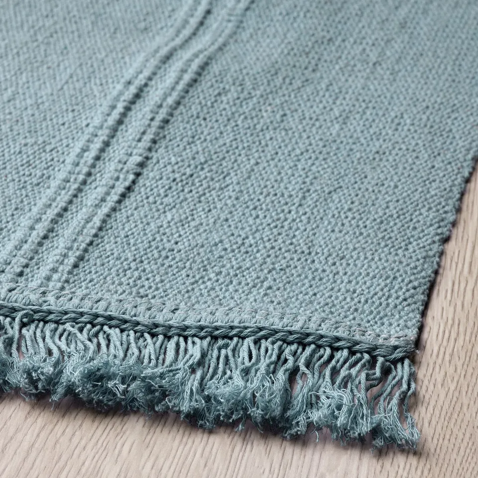Light blue woven rug with textured fringe lying on wooden floor. Durable and reversible, showcasing quality craftsmanship.
