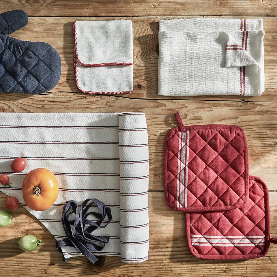 Image shows kitchen accessories: blue oven mitt, red potholders, white towels, rolled kitchen towel, tomatoes, onions, on rustic wood background.
