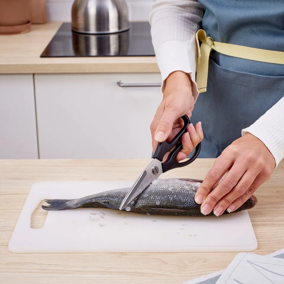 Person cuts fish with black kitchen scissors on cutting board.