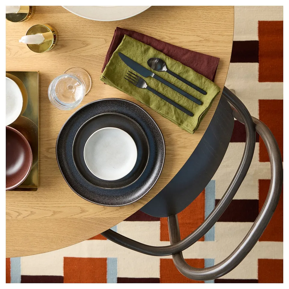 Top view of set table with black plate, green napkin, cutlery, glass, on patterned rug.