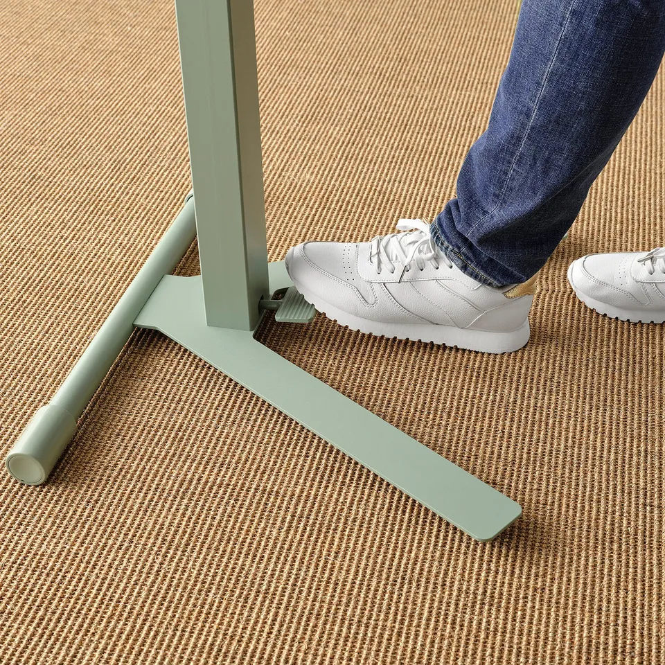Person using height-adjustable laptop table with light green metal base, white trainers, and patterned carpet.
