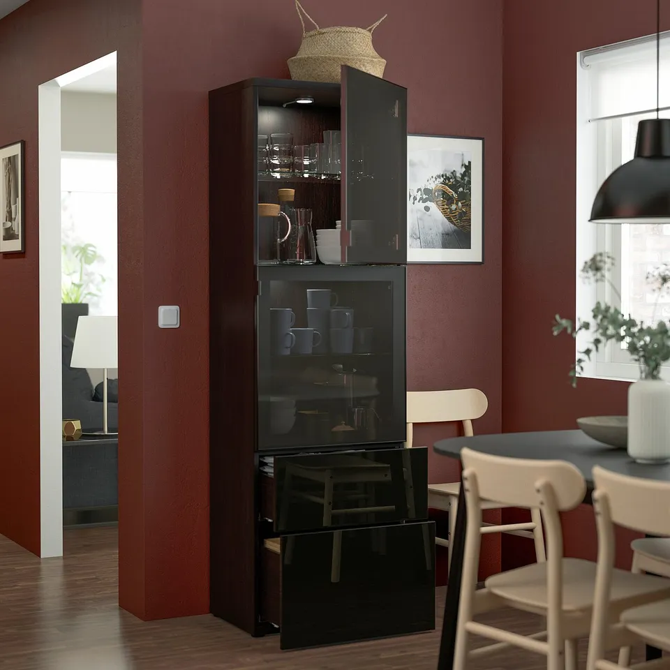 Black BESTÅ cabinet with glass doors and drawers stores and displays items. It's fixed to a red wall, showcasing cups and jars, under a woven basket.