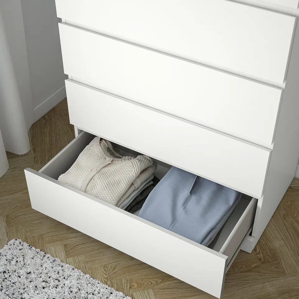 White chest of drawers with multiple drawers, one open showing neatly folded clothes. Safe design with wall anchoring.