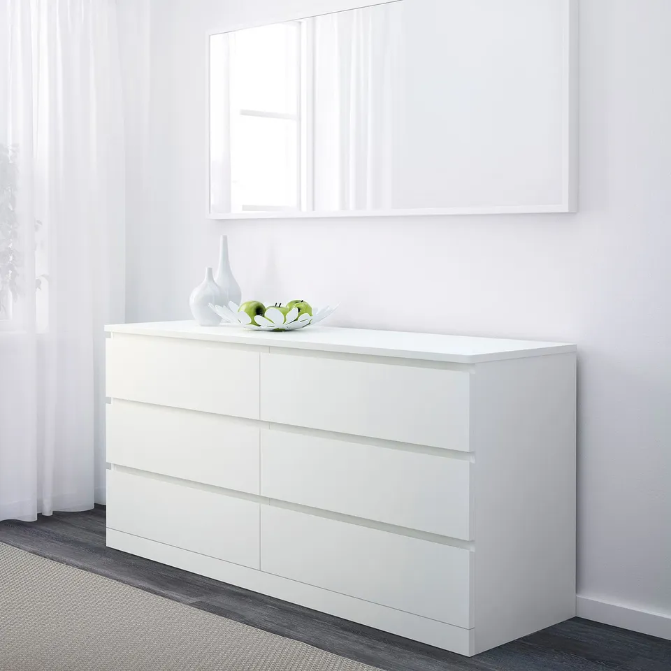 White chest with six drawers, smooth handles, topped with decorative bowl and fruit; positioned against white wall with mirror above.