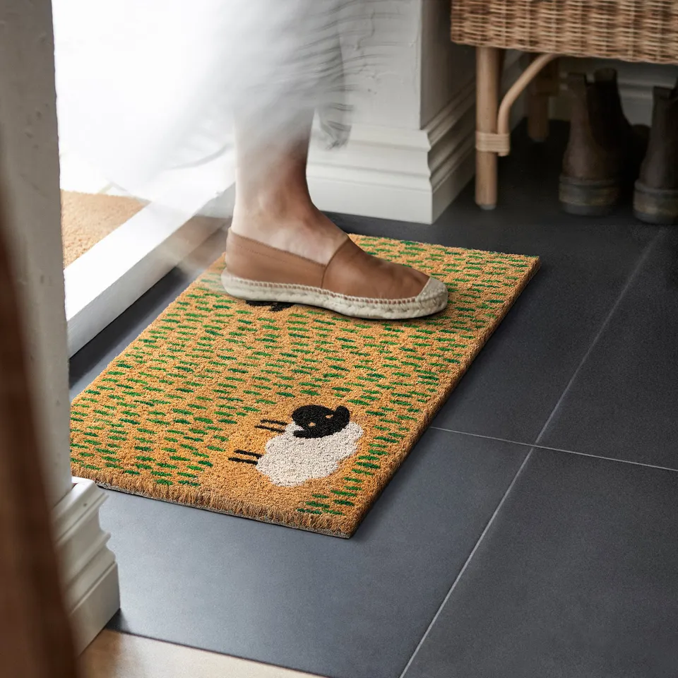 Person stepping on yellow-green door mat with sheep image, preventing dirt inside.