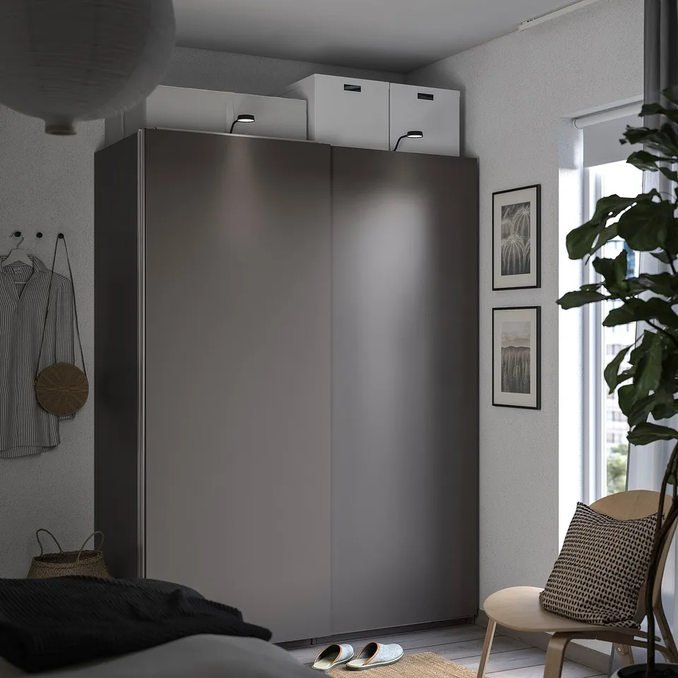 Modern bedroom with black PAX wardrobe, sliding doors, and white boxes on top. A chair with a cushion, plant, and framed pictures by the window.