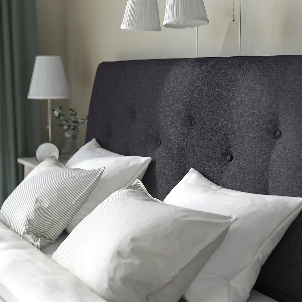 Upholstered bed with soft headboard, white pillows, and lamps. Slatted base for mattress support and ventilation.