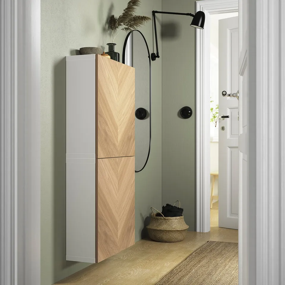Modern bathroom with tall, sleek light wood BESTÅ cabinet, black accent wall lamp, and wicker basket beside a white door.