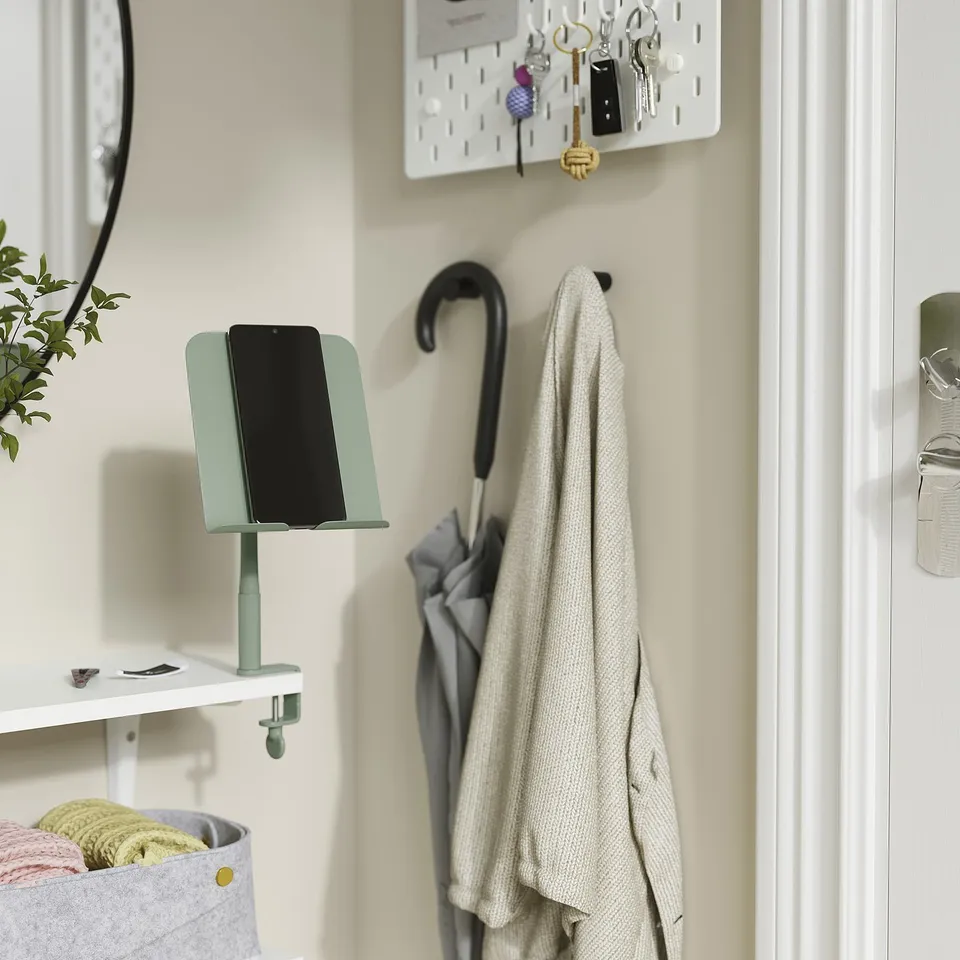 Organised entryway with RELATERA phone holder in green, clips on wall, holds black smartphone. Coats hang nearby on hooks.