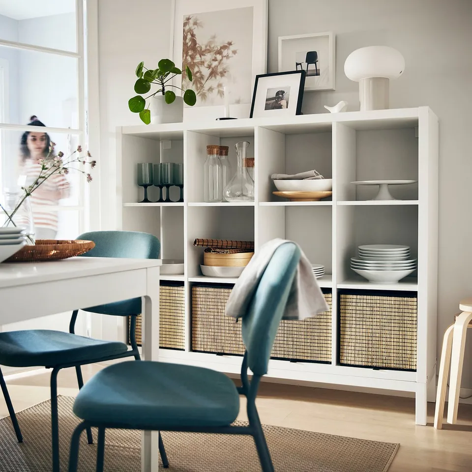 Modern dining area with white KALLAX shelf, blue cushioned chairs and various decorative items including glasses, plant, dishes, and framed photos.