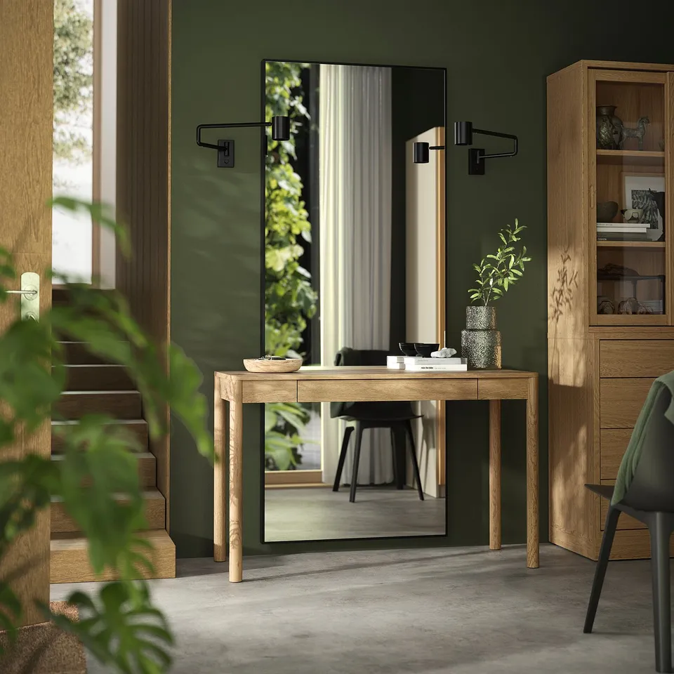 Natural wood console table between green walls and staircase, with drawer, slim legs, matching bookshelf, mirror, and green chair.