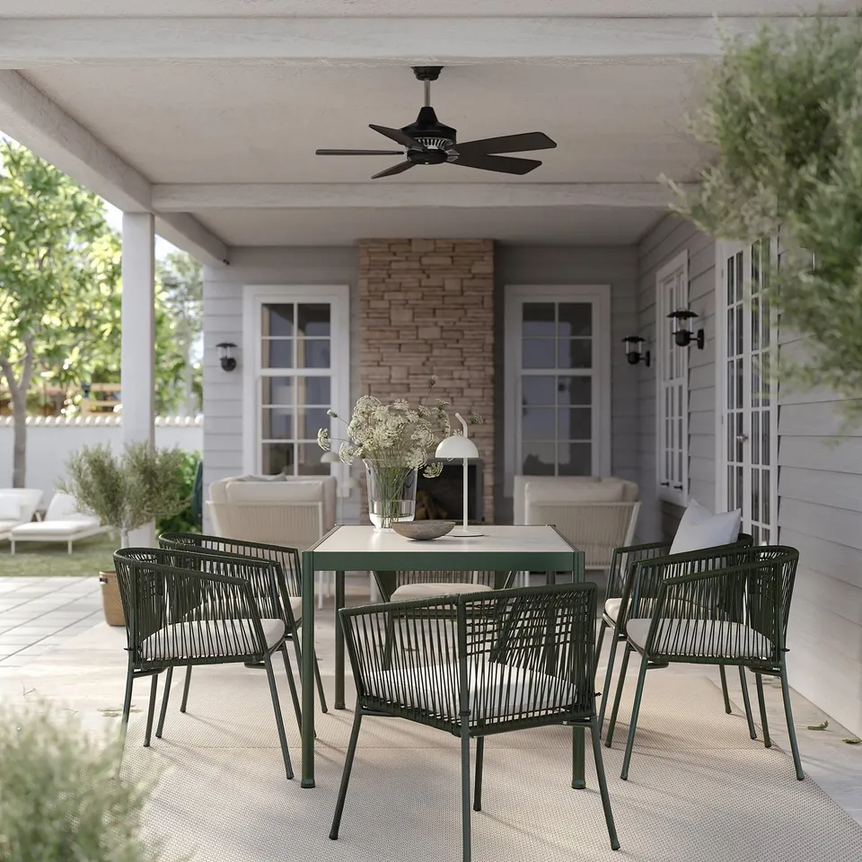 Outdoor seating area with dark green chairs and table, beige cushions, brick chimney, white ceiling fan, and flowers, on a patio.