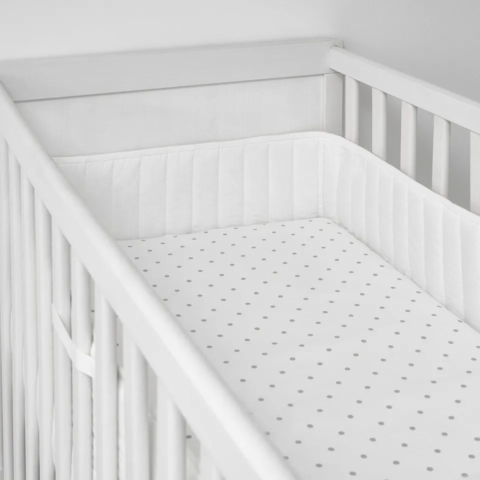 A white baby crib a padded bumper with discreet grey polka dots, safely securing a soft mattress inside.