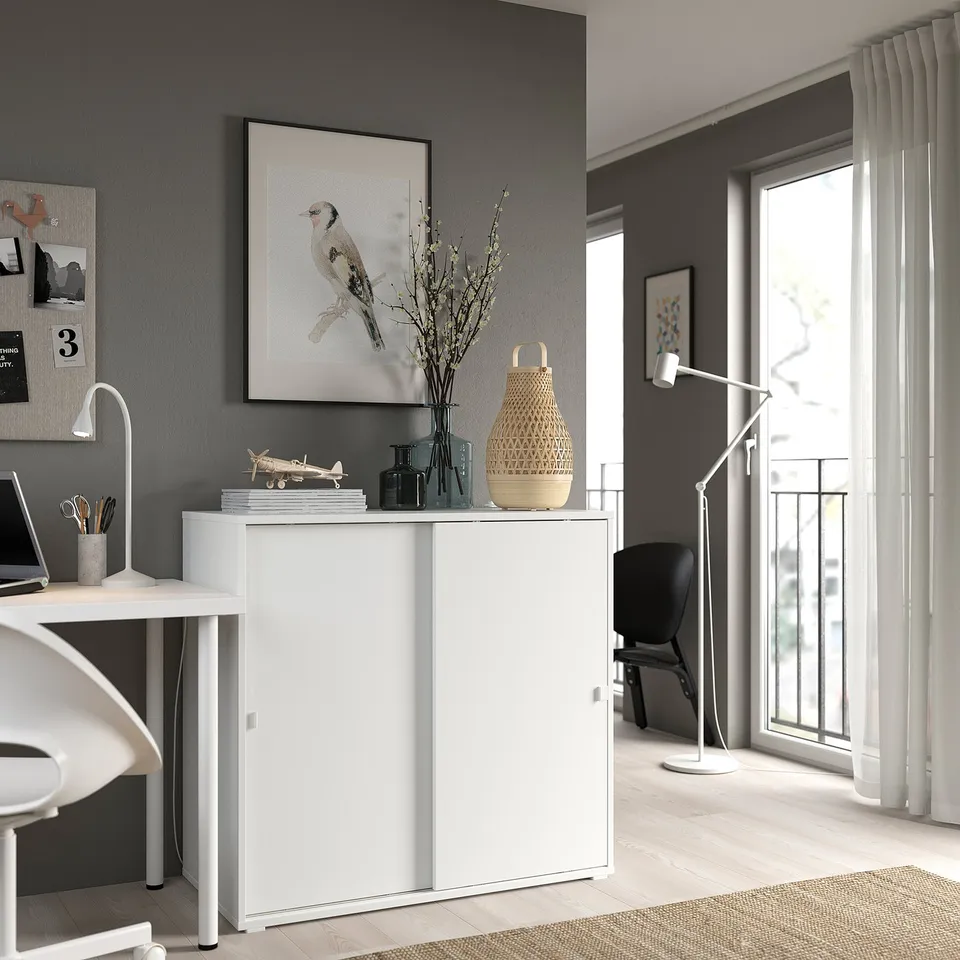 Modern home office with white IKEA VIHALS storage cabinet, sliding doors, grey walls, large window, and minimalist decor.