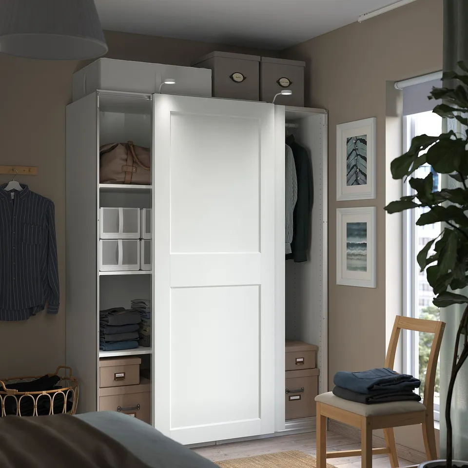 A modern, organised room with a white PAX wardrobe featuring sliding doors, housing folded clothes and hanging garments.