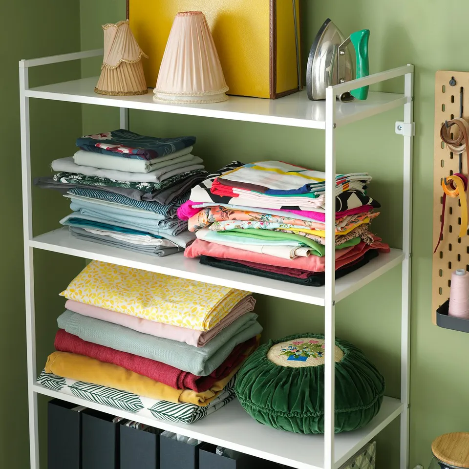 White 3-tier JONAXEL shelf holds colourful fabric stacks, decorative lamps, and iron in a green room.