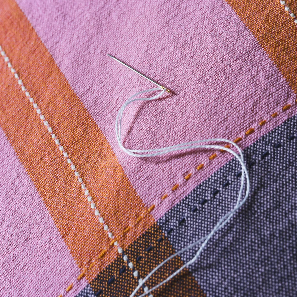 A close-up of handwoven fabric with pink, orange, and blue stripes showing fine stitching and needle with thread.