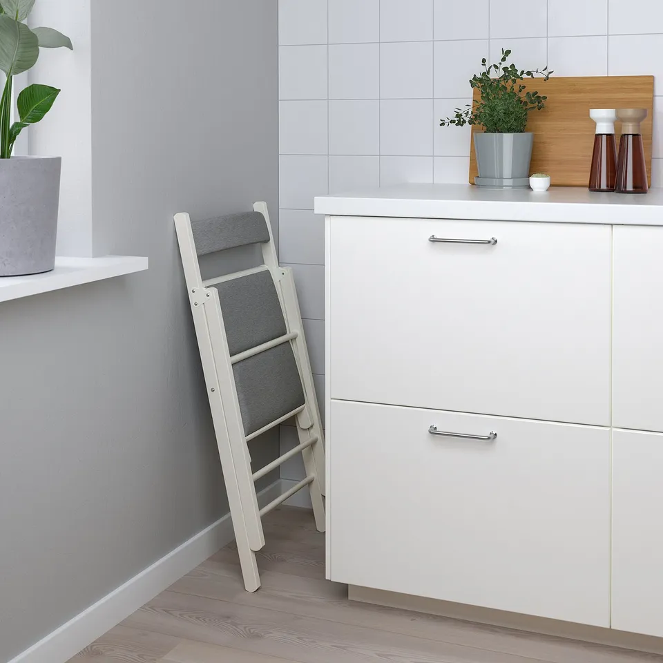 A white kitchen with a foldable chair, cabinet, and countertop decorations.