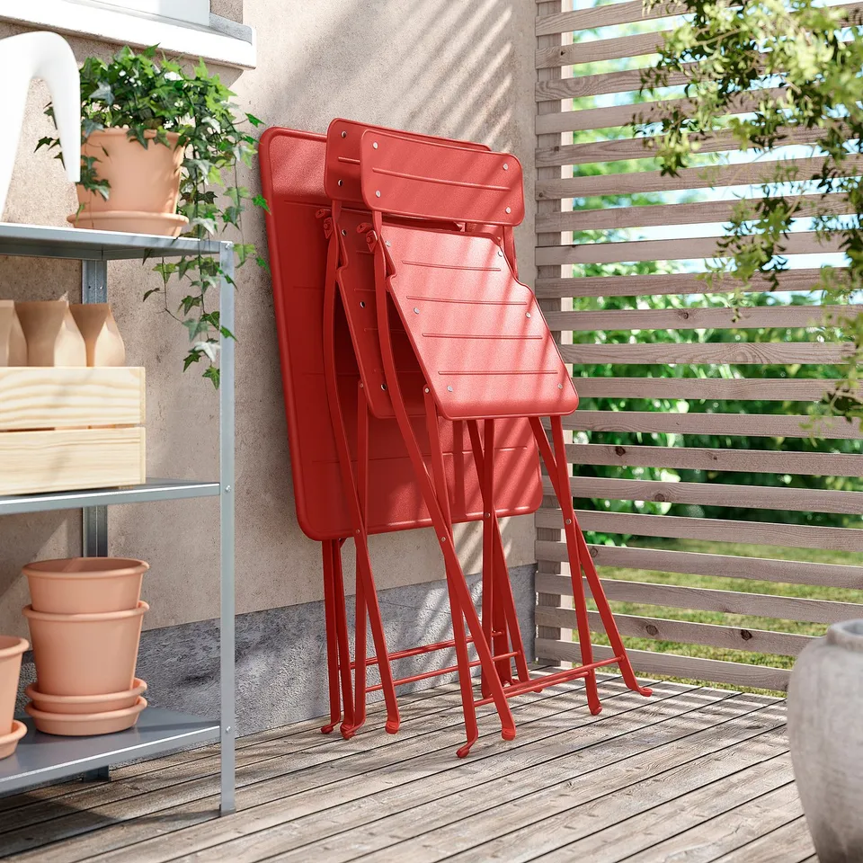 Three bright red SUNDSÖ chairs stacked neatly on a wooden patio next to potted plants and a metal shelf.