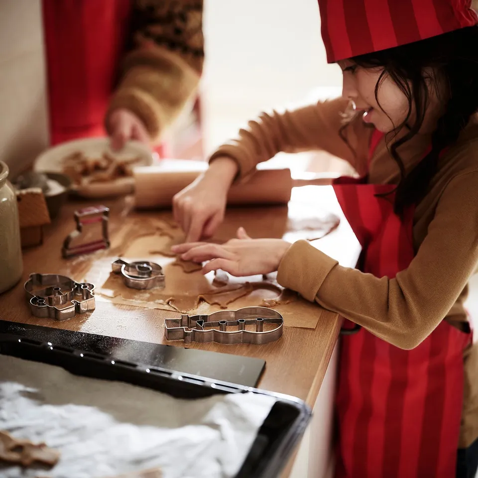 Child in red apron, baking cookies with festive cutters. Adjustable for ages 4+.