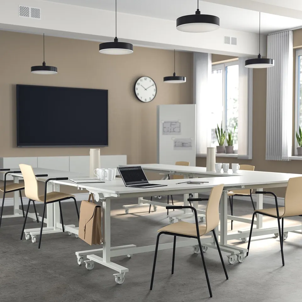 Modern office with MITTZON tables & läktare chairs. White foldable tables, beige stackable chairs, black pendant lights, large screen & clock.