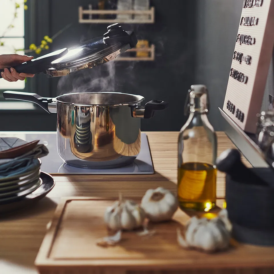 A person is lifting a lid from a steaming IKEA 365+ pressure cooker on a wooden countertop. Nearby are garlic, olive oil, plates, and a menu board.