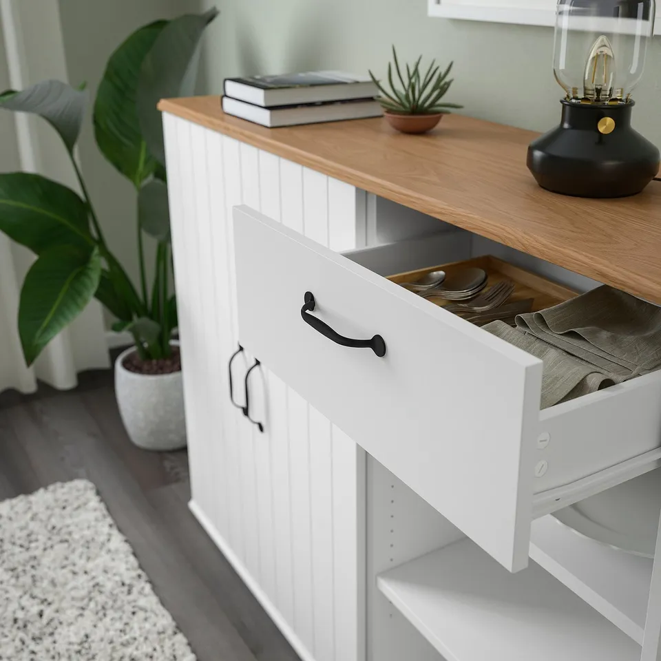 A wooden-topped white cabinet with an open drawer revealing dishes and utensils. Nearby, a large green plant and a stack of books on the cabinet.
