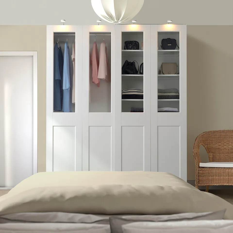 White PAX wardrobe with glass panels, beside bed, showcasing clothes and accessories.