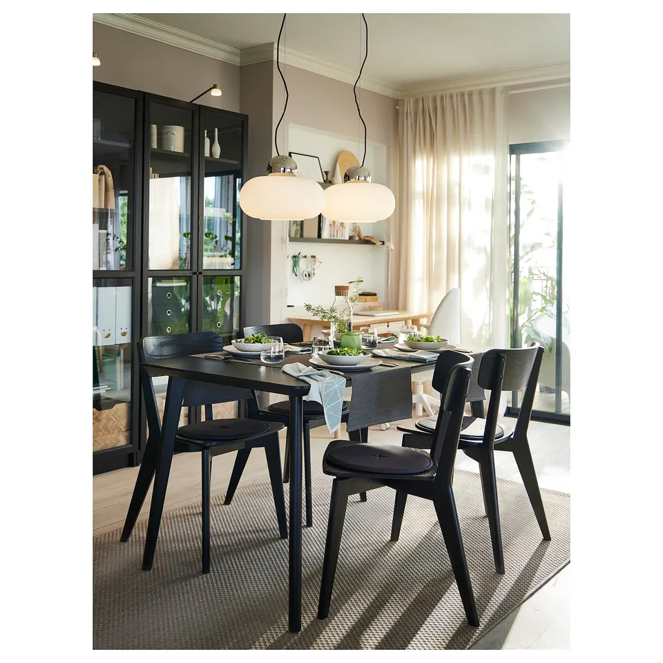 Black dining table with 4 chairs, plates, greenery, under modern light in cosy room with large windows and rug.