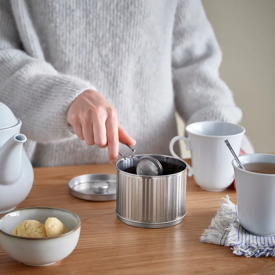 Person pours tea into silver BETTHAJ tin on wooden table with mugs and cookies.