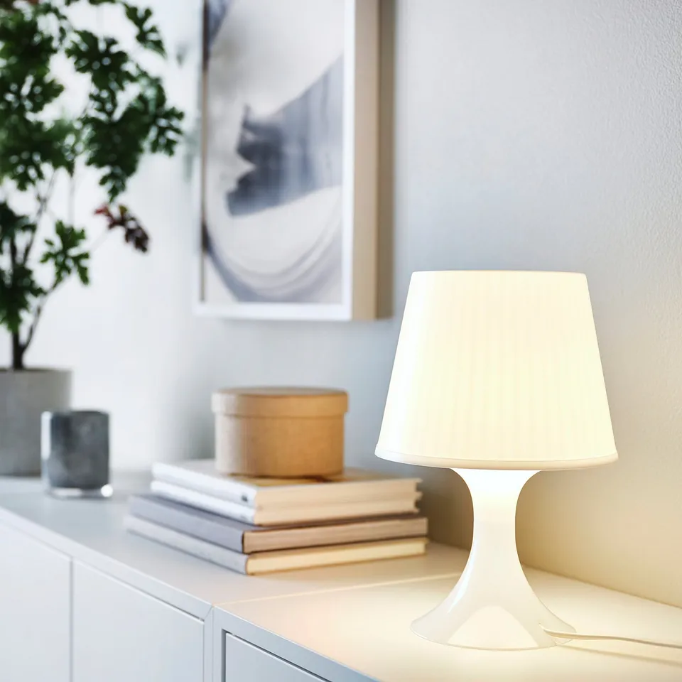 A white LAMPAN table lamp with a curved design sits on a surface near books and a plant. Its small size complements the minimalist decor.