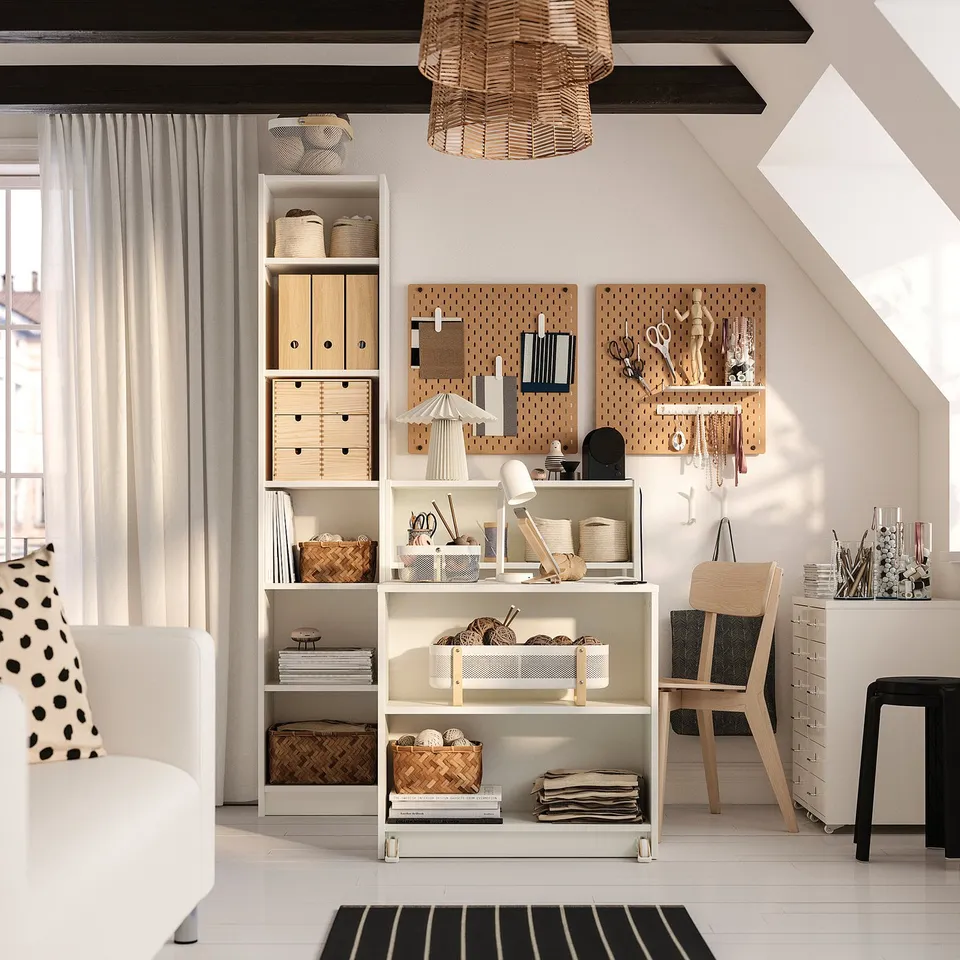 White BILLY bookcase combo with foldable table, open shelves, and various items next to sofa and chair, lit by table lamp and pendant light.