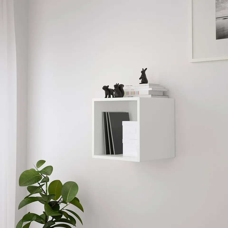 White wall-mounted cabinet EKET with open storage, holding various items including books, black figurines, and folders. Greenery at bottom left.