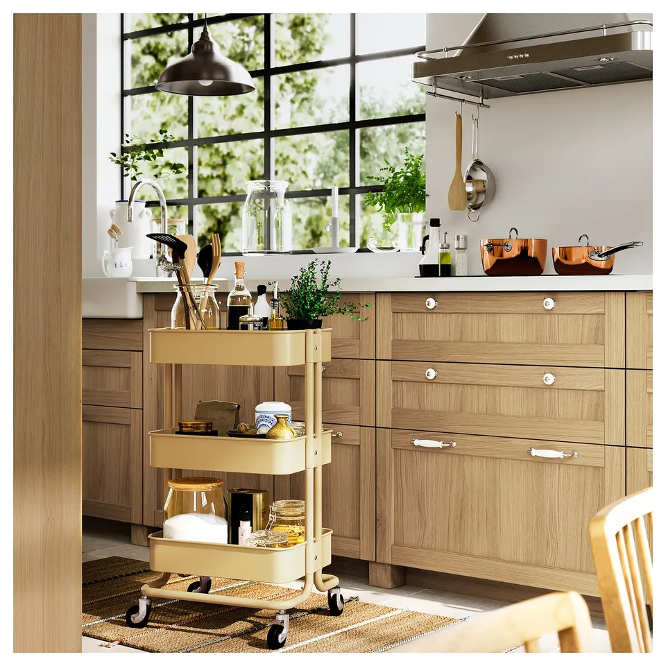 Modern kitchen with wooden cabinets, a cream-colored rolling cart stocked with kitchen items, a large window, and hanging utensils.