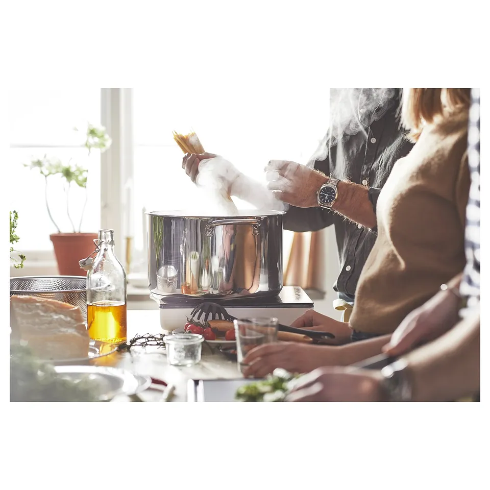 Two people cooking with IKEA 365+ pots. One holds a steaming lid; counter has food and utensils.