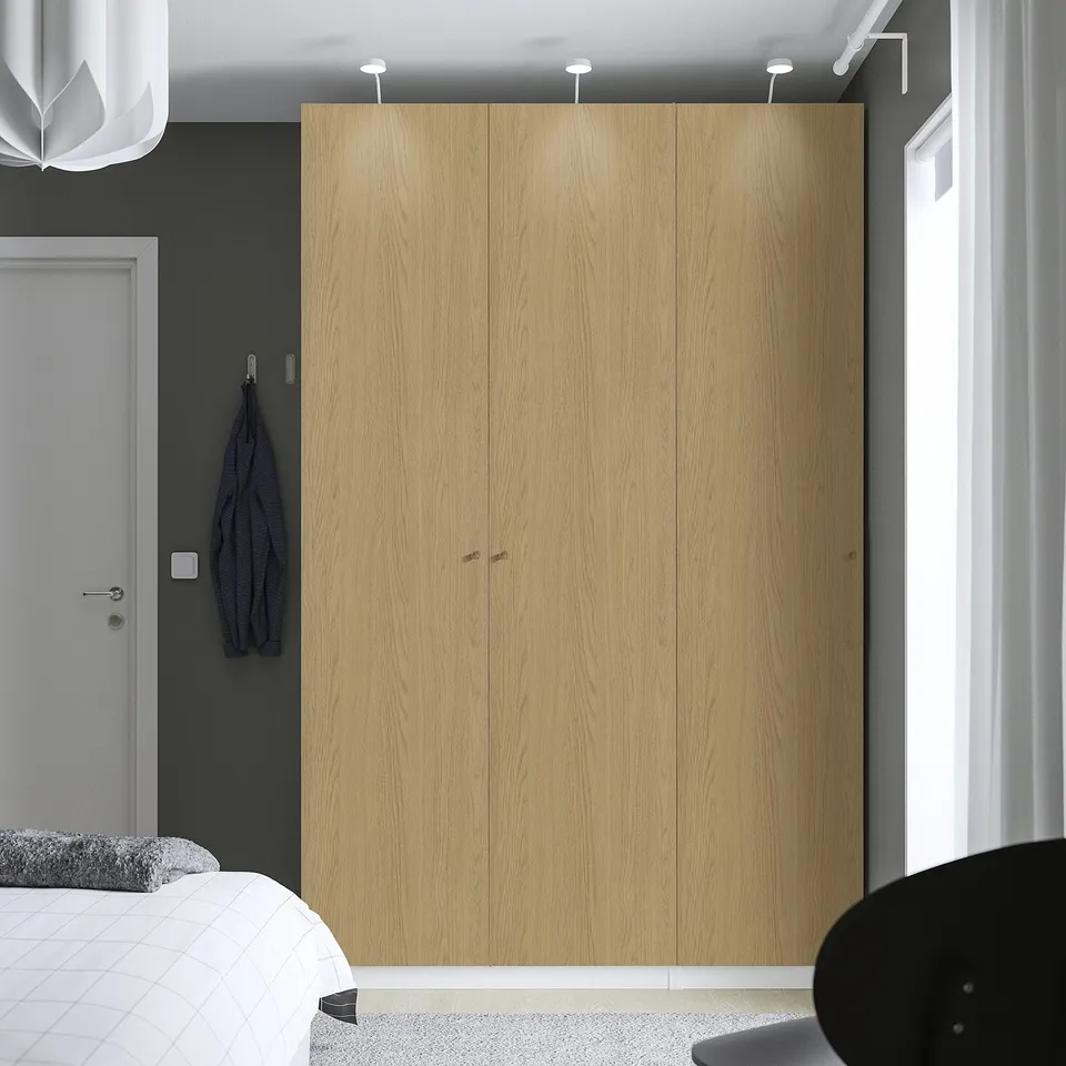 Modern bedroom with large light wood PAX wardrobe, grey wall, hanging jacket, and partially visible bed.